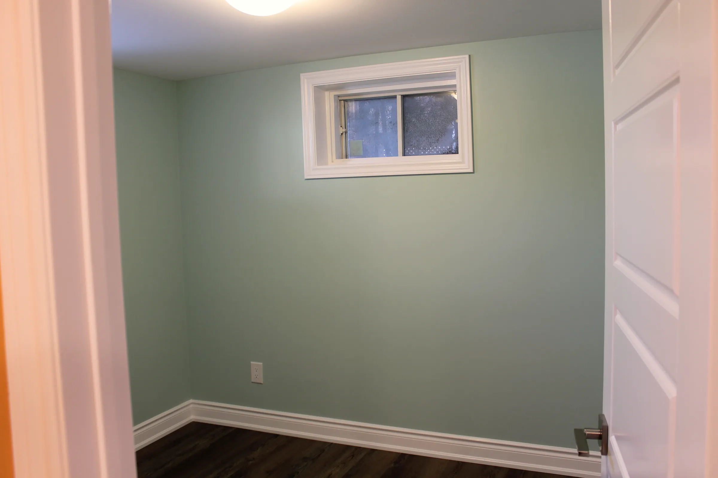 Finished basement bedroom with green walls, a hopper window, white trim, and dark wood-look floor.