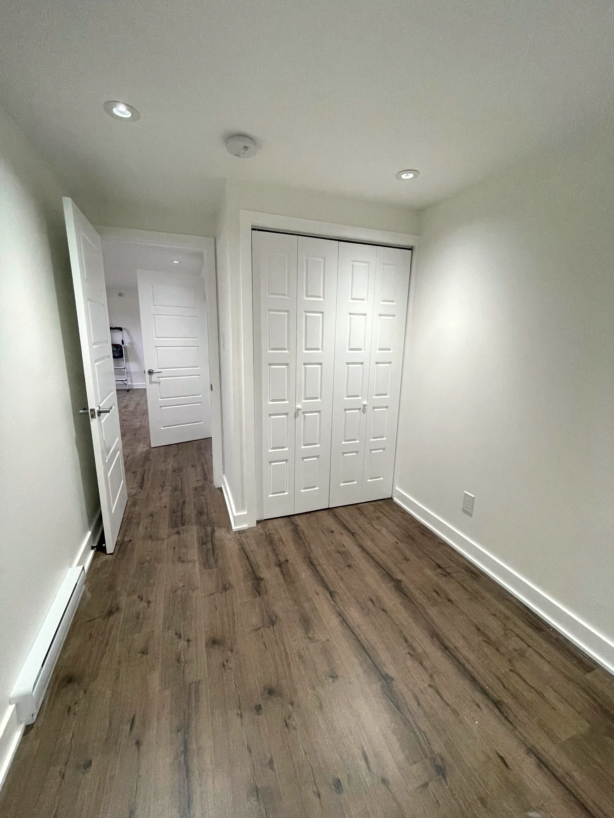 Finished basement bedroom with white folding closet doors, baseboard heater, wood-look plank floor, and an open door to the hallway.