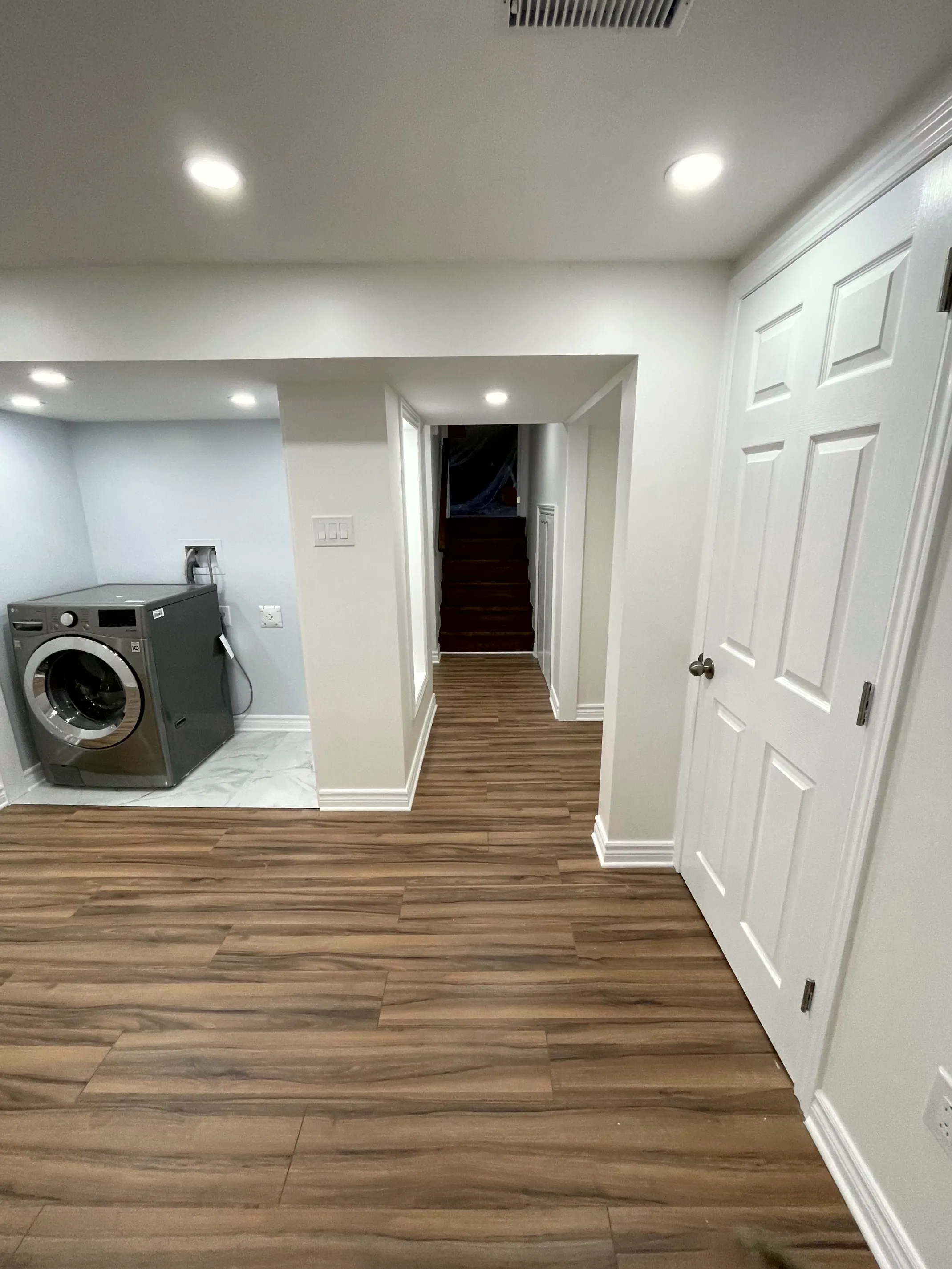 Finished basement with wood-look plank floor, recessed lighting, a side laundry alcove with washer, and a hallway through to the stairs.