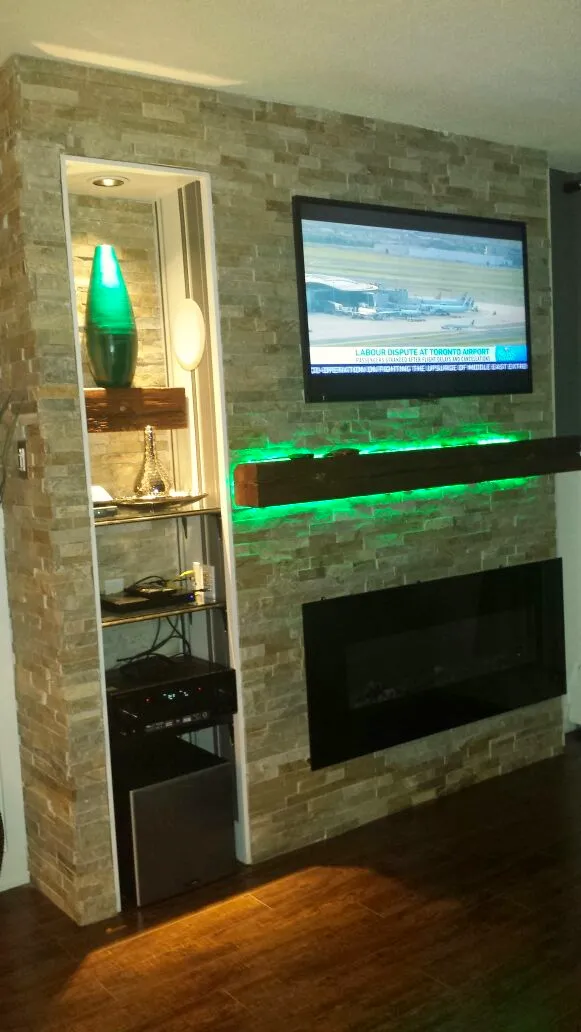 Finished basement media wall with stacked-stone surround, mounted TV, wood floating shelf with LED accent, and a linear electric fireplace.