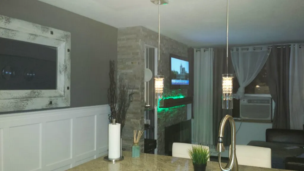 Wide finished basement with stone fireplace wall, white panelled wainscoting, a stone-look kitchenette counter, and two pendant lights.