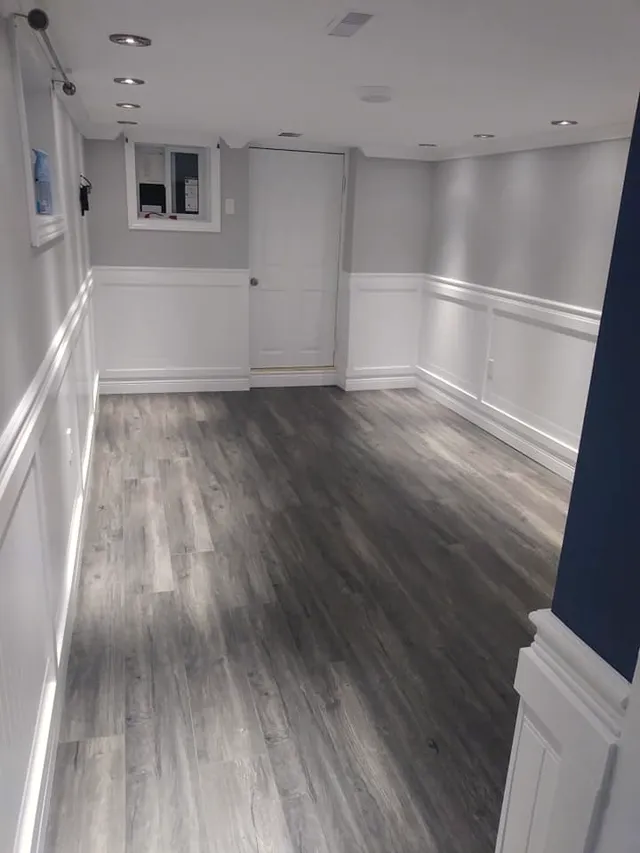 Finished basement room with grey wood-look plank floor, white wainscoting, recessed lighting, and an electrical panel set in a wall niche.
