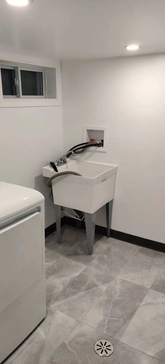 Finished basement laundry area with stone-look floor tile, white utility sink on metal legs, recessed washer hookup box, and a floor drain.