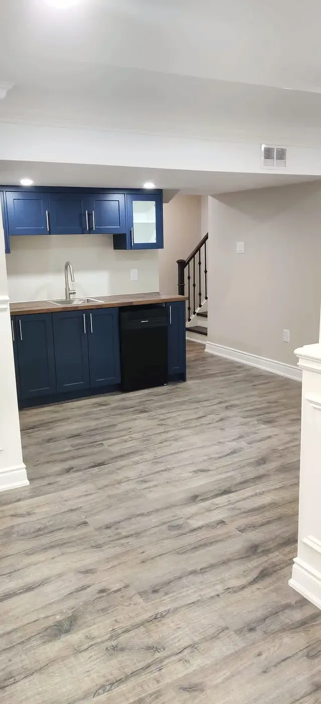 Finished basement wet bar with navy shaker cabinets, butcher-block counter, bar fridge, and a staircase with black railing.