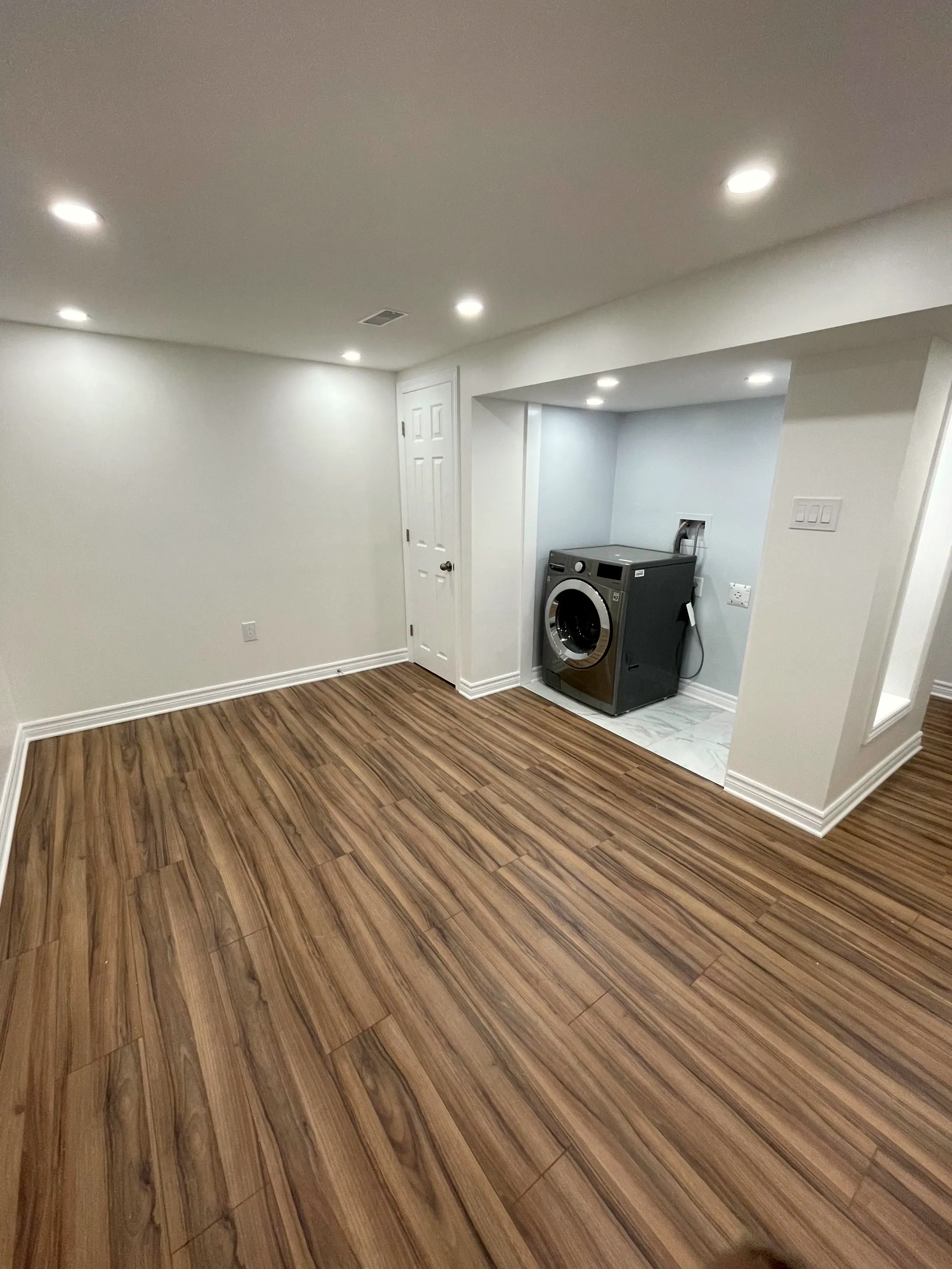 Finished basement room with wood-look floor, recessed lighting, and a side laundry alcove with a front-load washer.
