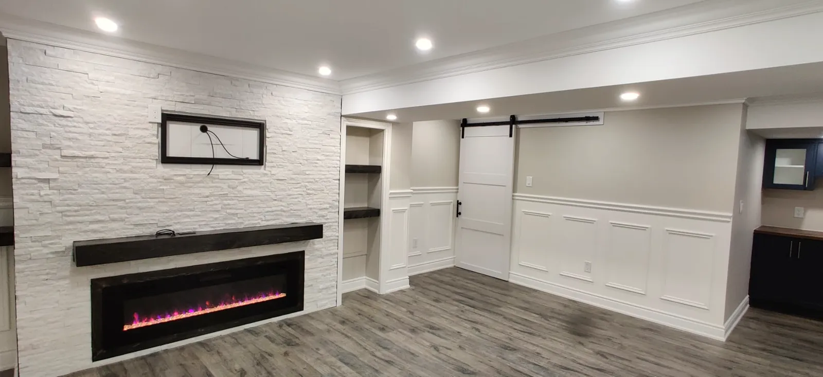 Finished basement family room with a stone-look fireplace wall, electric fireplace, built-in shelving, wainscoting, and a sliding barn door.