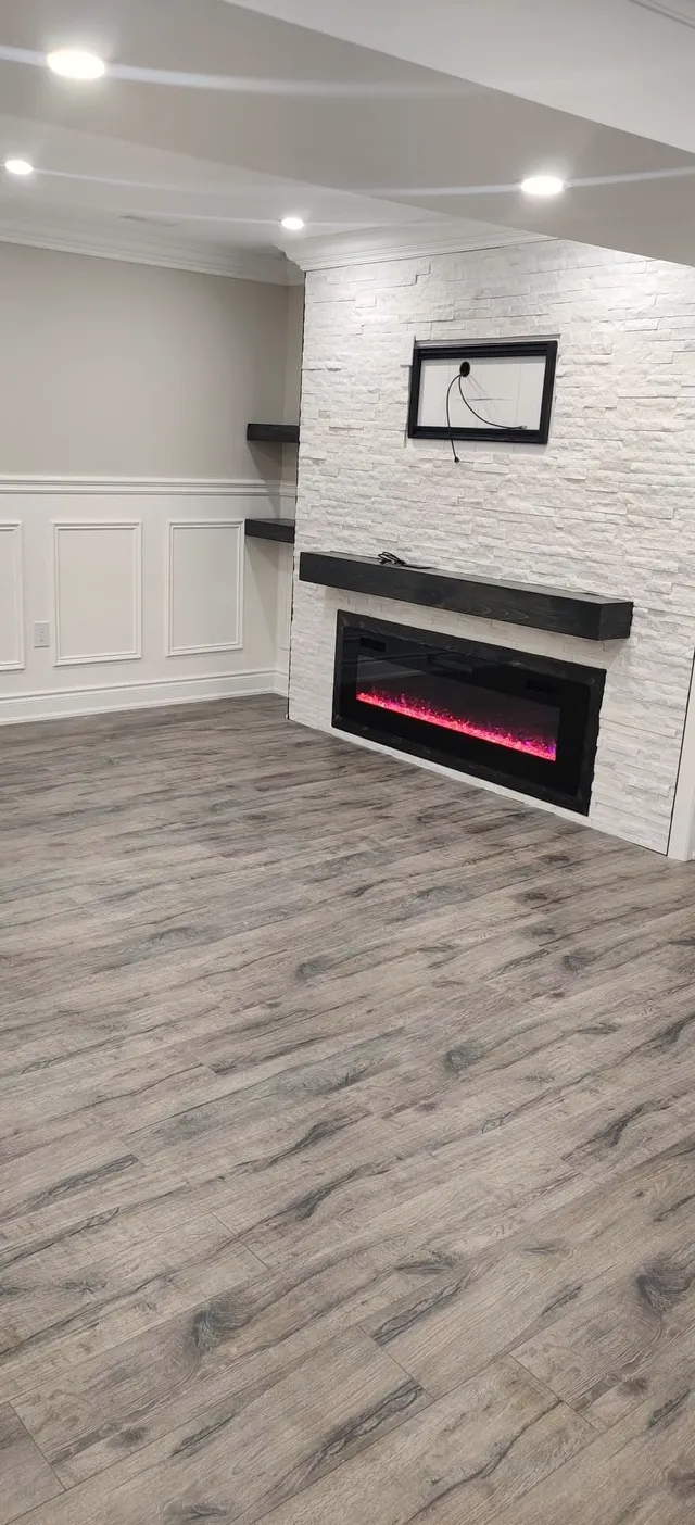 Finished basement family room with a stone-look fireplace wall, electric fireplace, floating shelves, wainscoting, and wood-look floor.