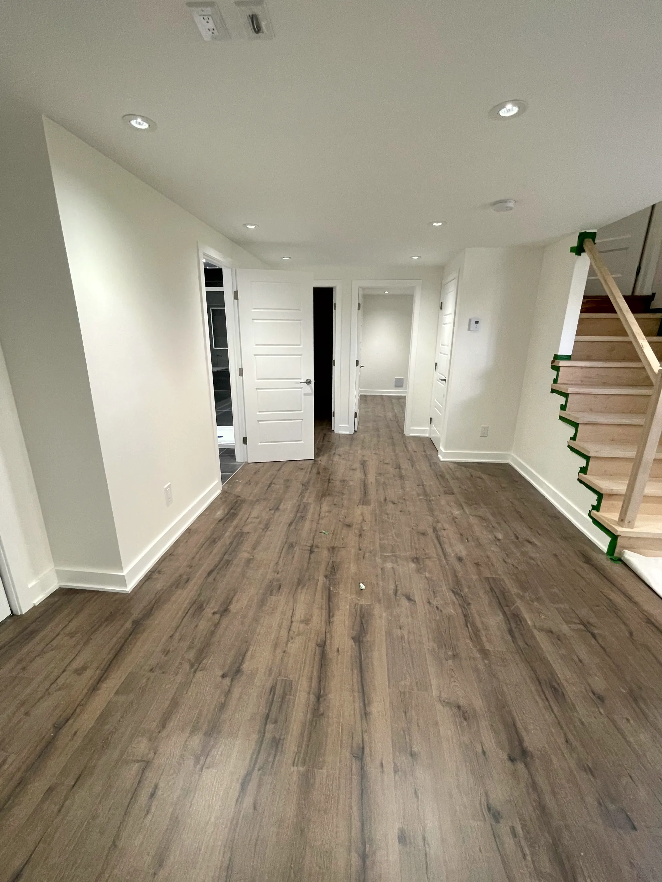 Finished open basement with wood-look floor, recessed lighting, several white doors, and a new staircase taped for finishing.