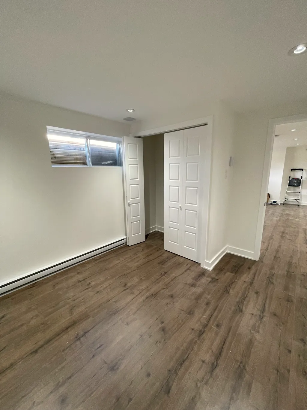 Finished basement bedroom with a hopper window, bi-fold closet door, electric baseboard heater, and wood-look floor.