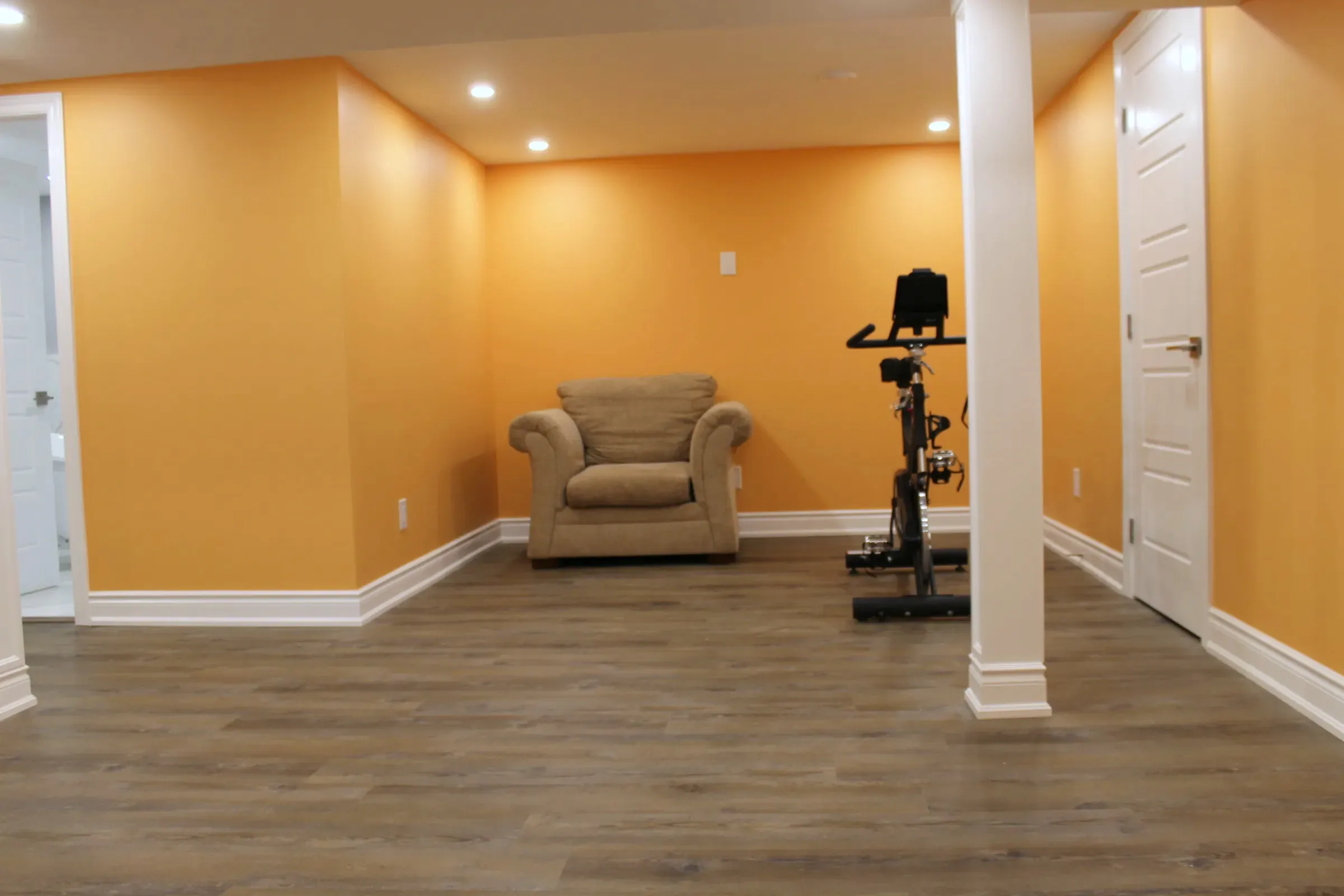 Finished basement room painted orange, with a recliner, a stationary bike, a support post, and wood-look floor.