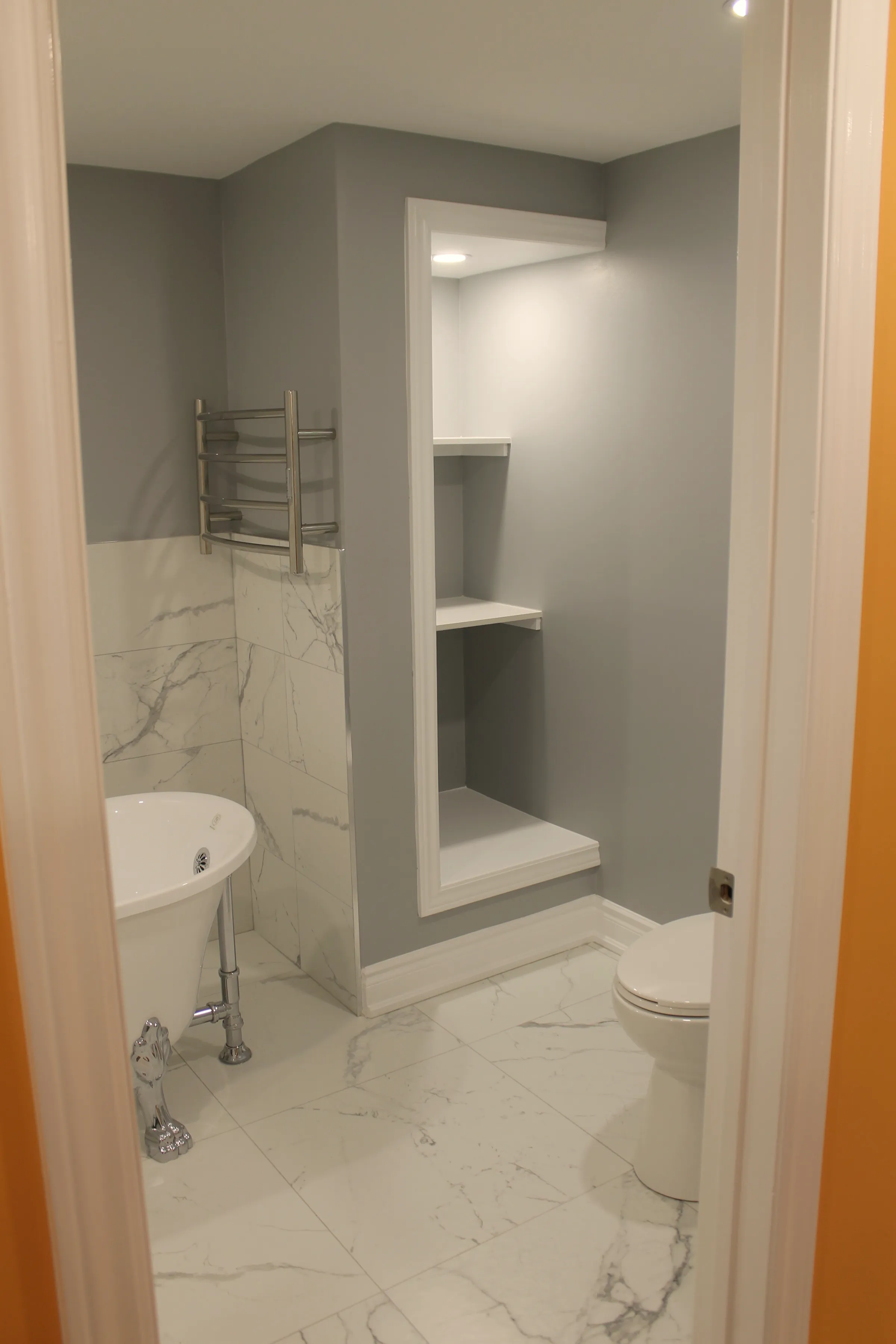 Finished bathroom seen from the doorway, freestanding tub on chrome feet, chrome towel warmer and a recessed niche shelf.
