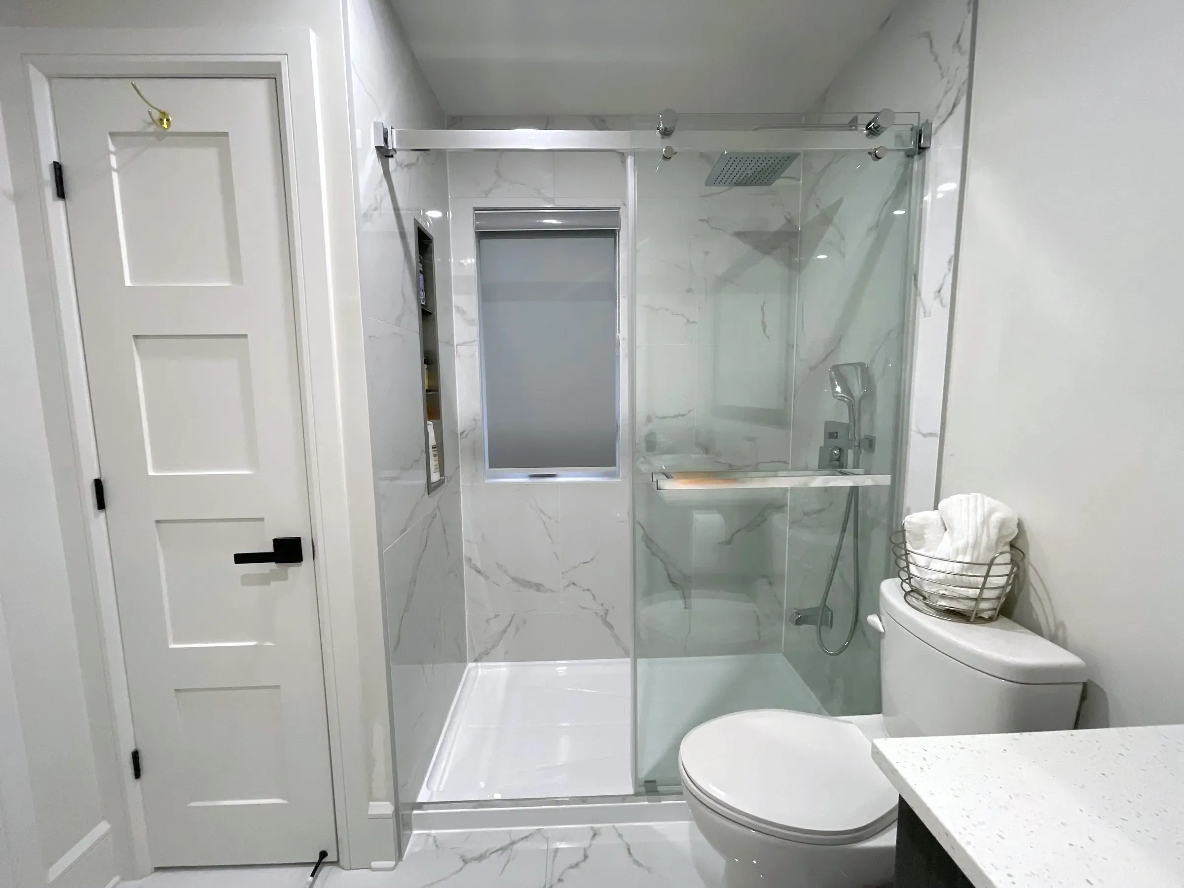 Finished bathroom, glass shower with sliding door, stone-look wall tile, recessed shelf, toilet and white vanity counter.