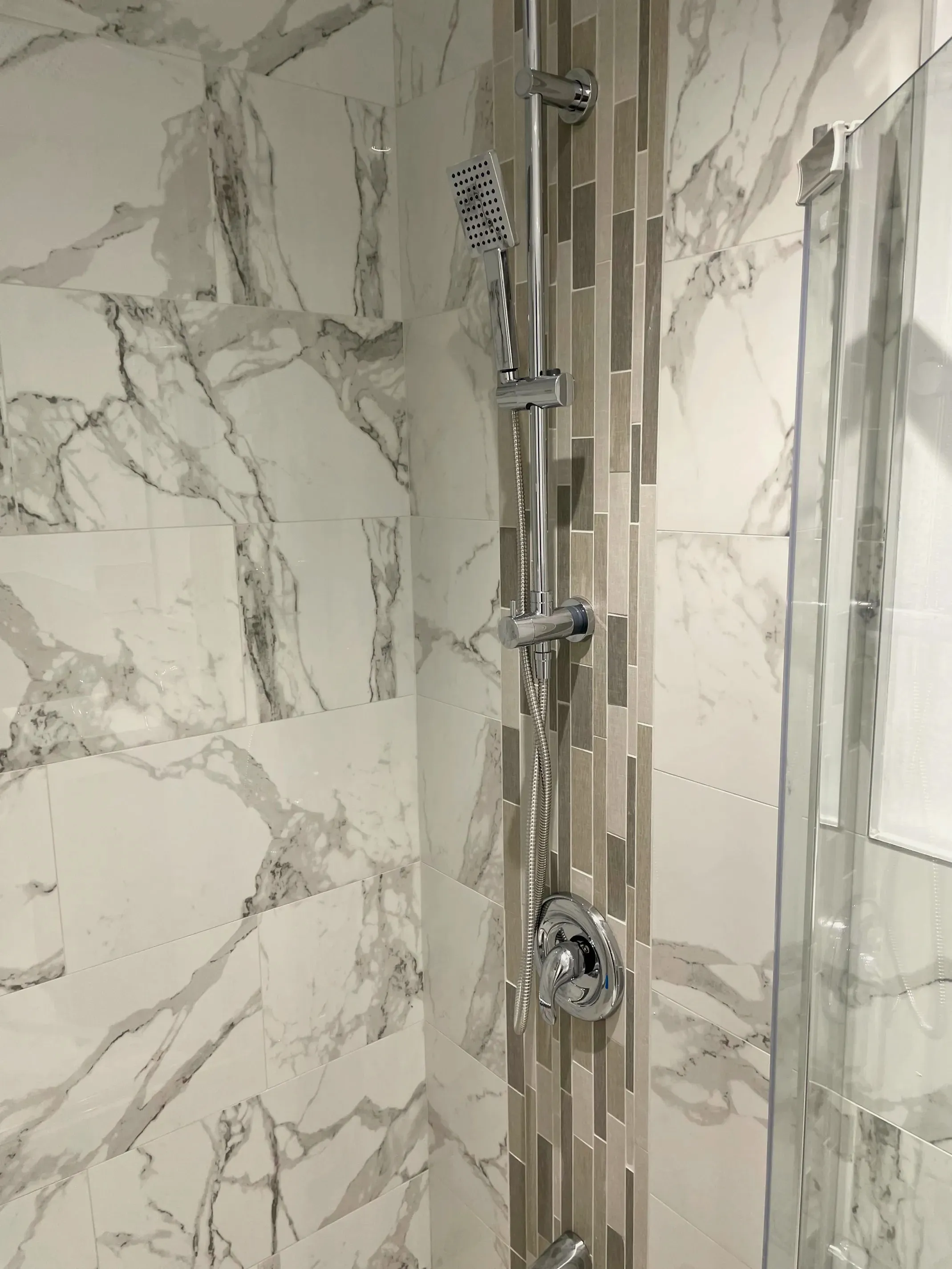 Finished shower wall in stone-look tile with a chrome slide-bar handheld and a vertical mosaic accent strip.