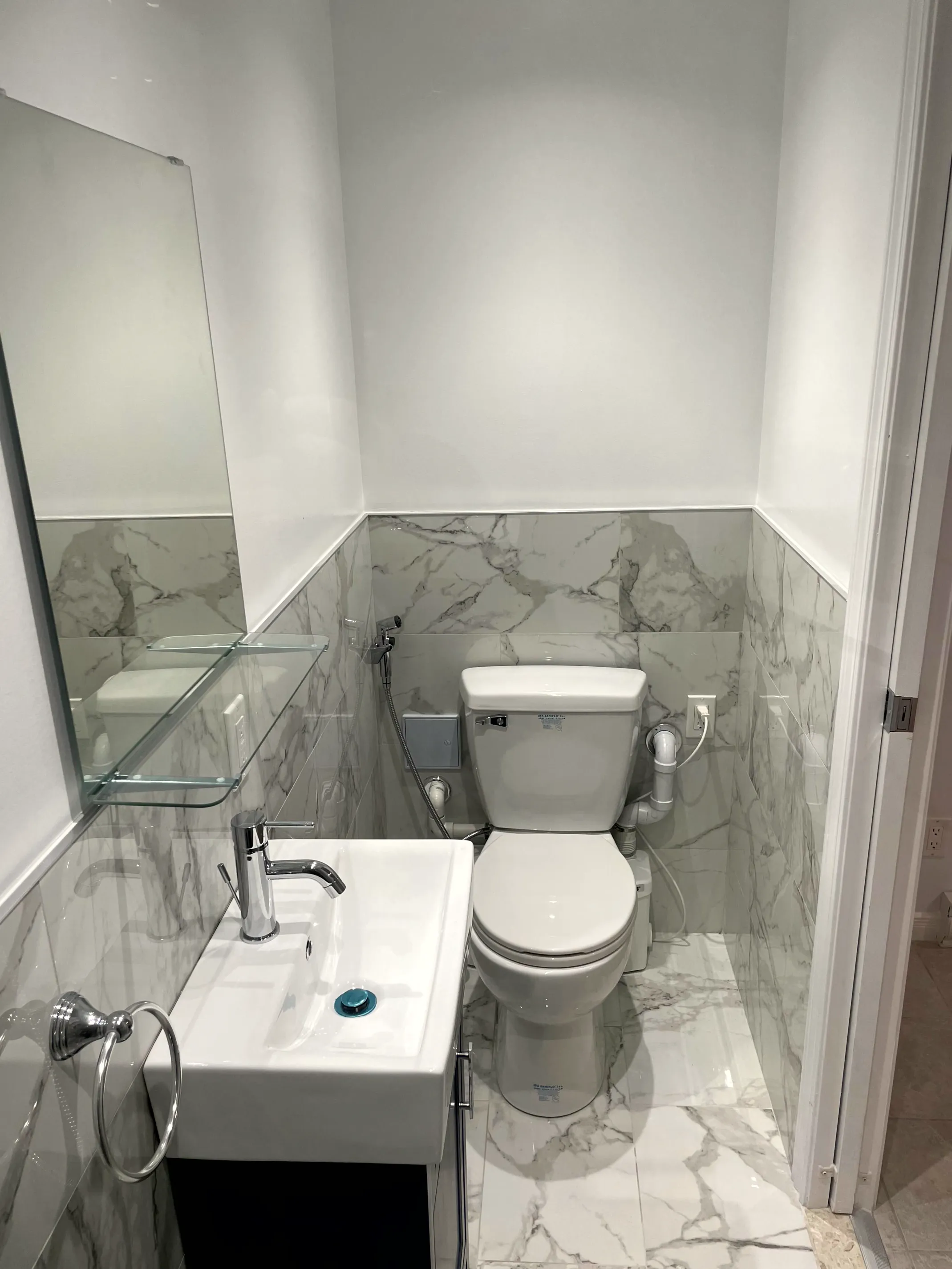 Finished half bath, stone-look wall tile, white wall-hung sink, tall mirror, glass shelf and toilet with hand bidet sprayer.