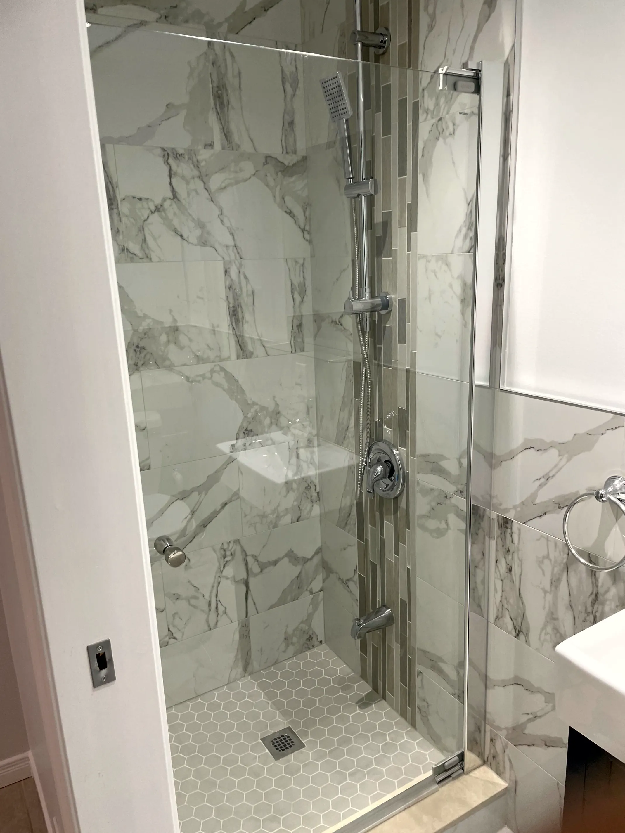 Finished walk-in shower, glass door, stone-look wall tile, vertical mosaic accent strip, corner shelf and white hex floor.