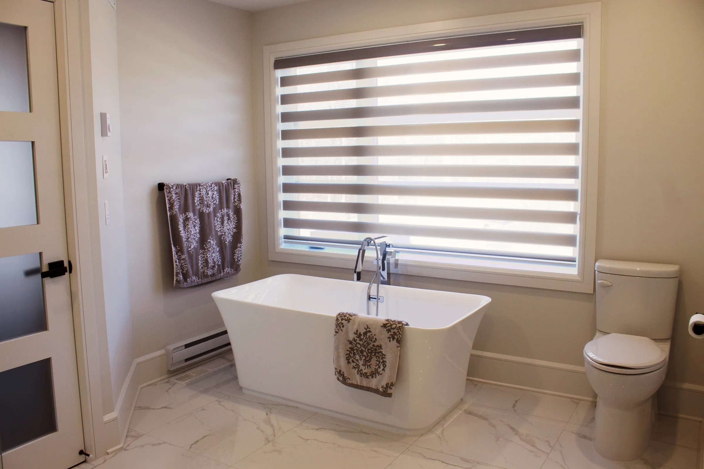 Finished bathroom, white freestanding tub under a wide window with zebra shade, baseboard heater, light stone-look floor.