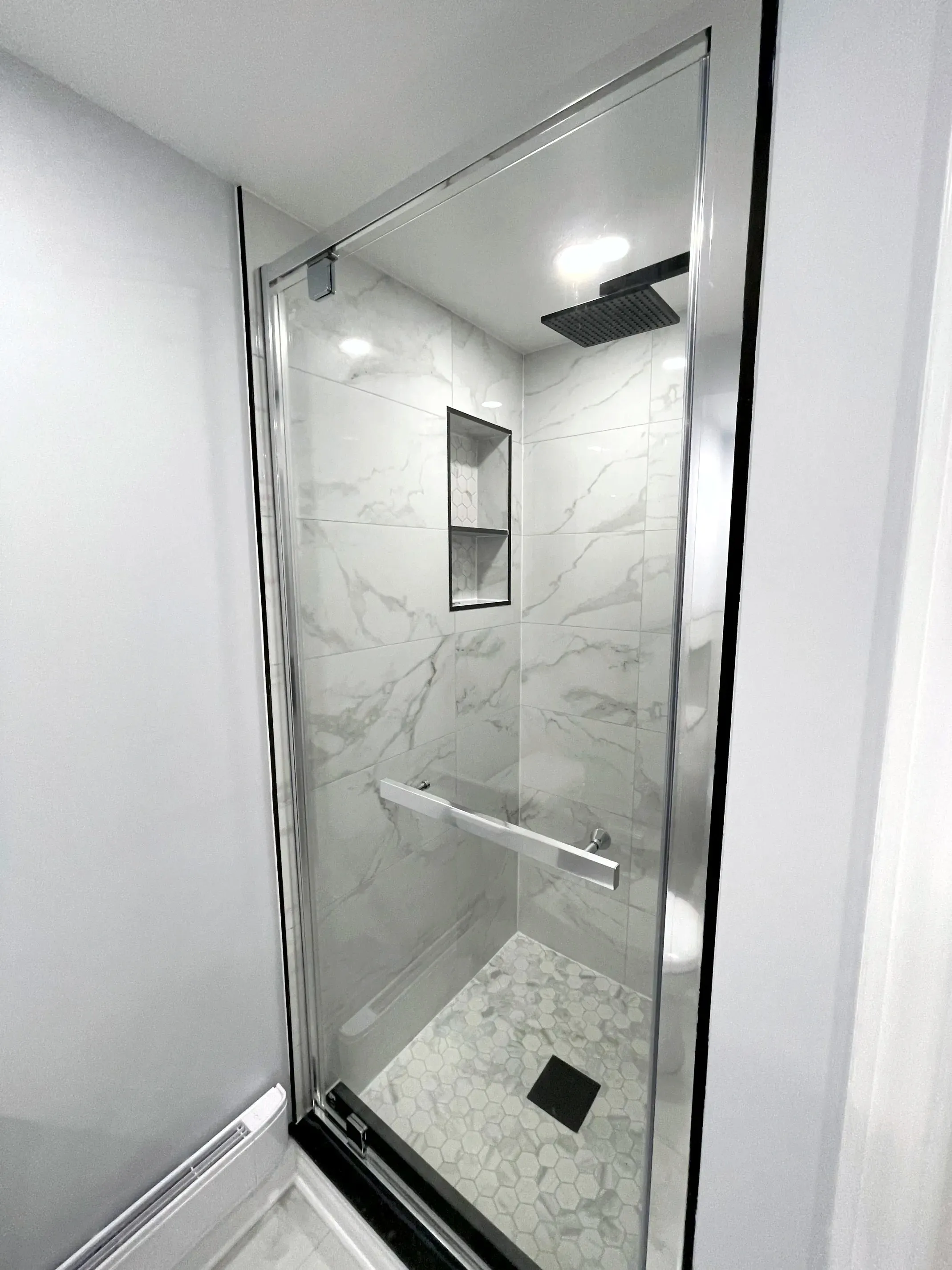 Finished walk-in shower, frameless glass door, light stone-look wall tile, rainhead, recessed niche, mosaic floor, grab bar.