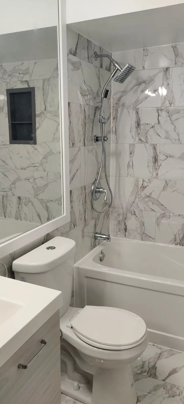 Finished tub and shower combo with stone-look wall tile, recessed niche, chrome rainhead and handheld, and white vanity.