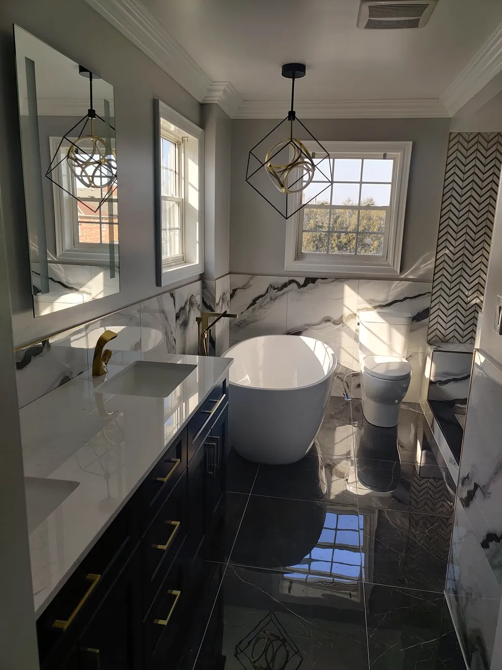 Finished bathroom, dark double vanity with gold faucets, white freestanding tub, two geometric pendants, dark stone-look floor.
