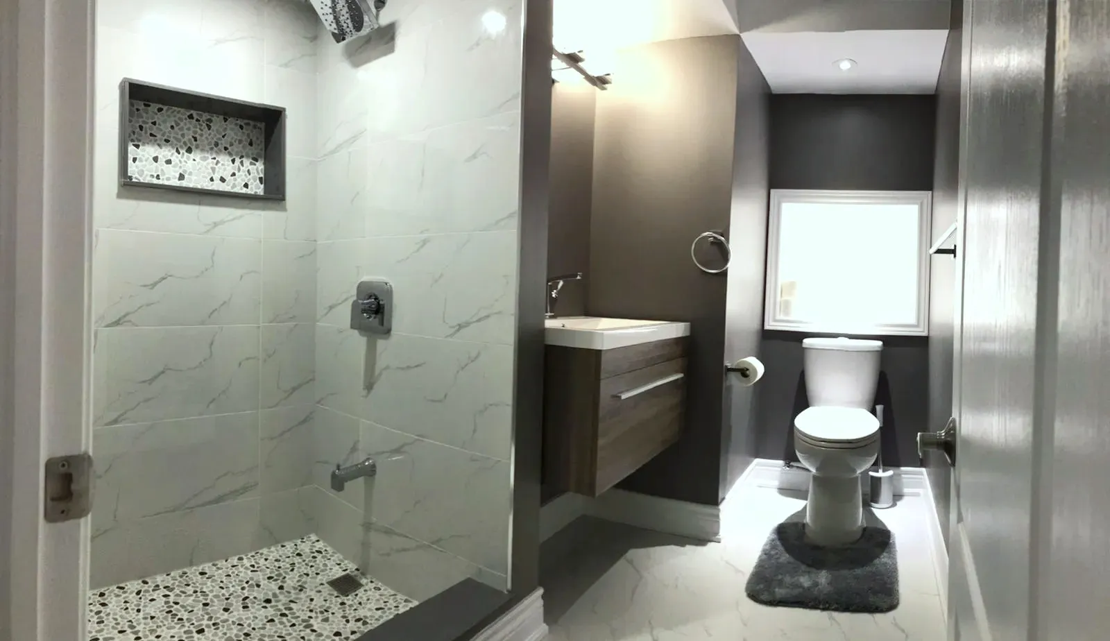 Finished bathroom, walk-in shower in light stone-look tile, pebble-mosaic floor, recessed niche, floating wood-look vanity, dark accent wall.