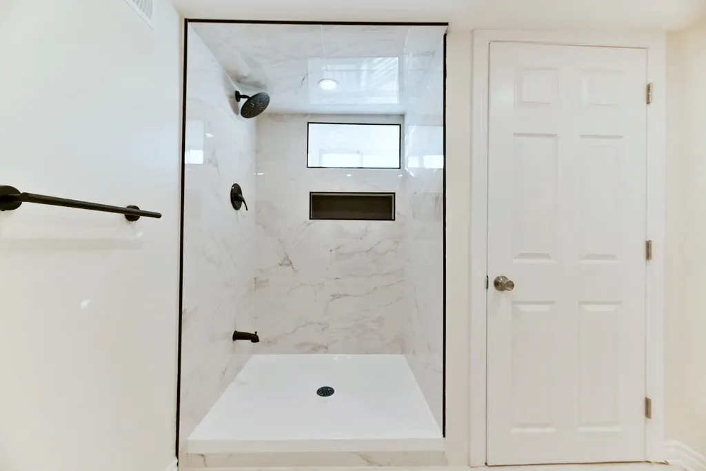 Finished walk-in shower alcove, white stone-look wall tile, black fixtures, recessed niche, transom window, white shower base.