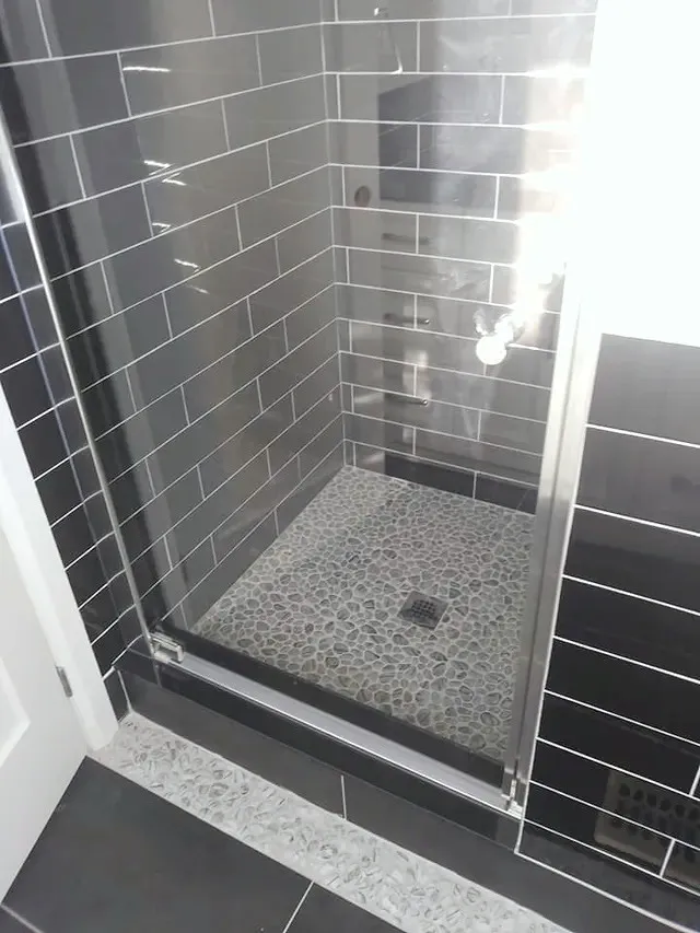 Finished walk-in shower, dark glossy wall tile, pebble-mosaic floor, frameless glass door, dark floor tile.