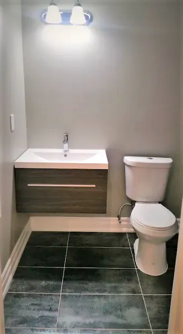 Finished powder room, floating wood-look vanity, integrated white sink, chrome faucet, vanity light, toilet, dark stone-look floor.