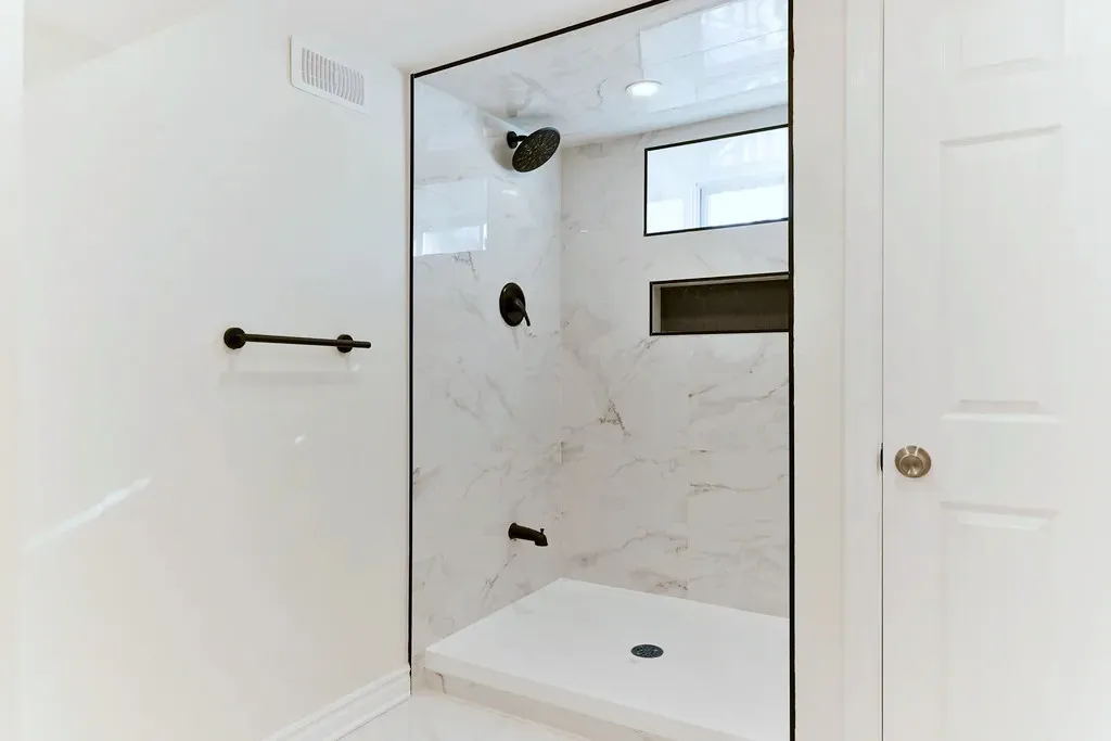Finished shower stall with light stone-look wall tile, black trim, matte black rainhead, recessed niche, and a window above.