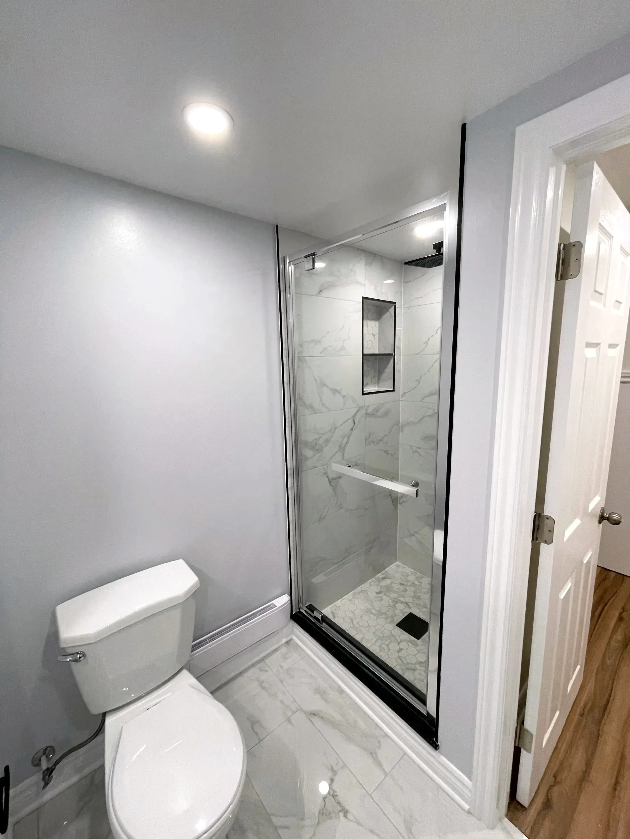 Finished basement bathroom with a glass shower in stone-look tile, recessed niche, toilet, and wood-look floor beyond.