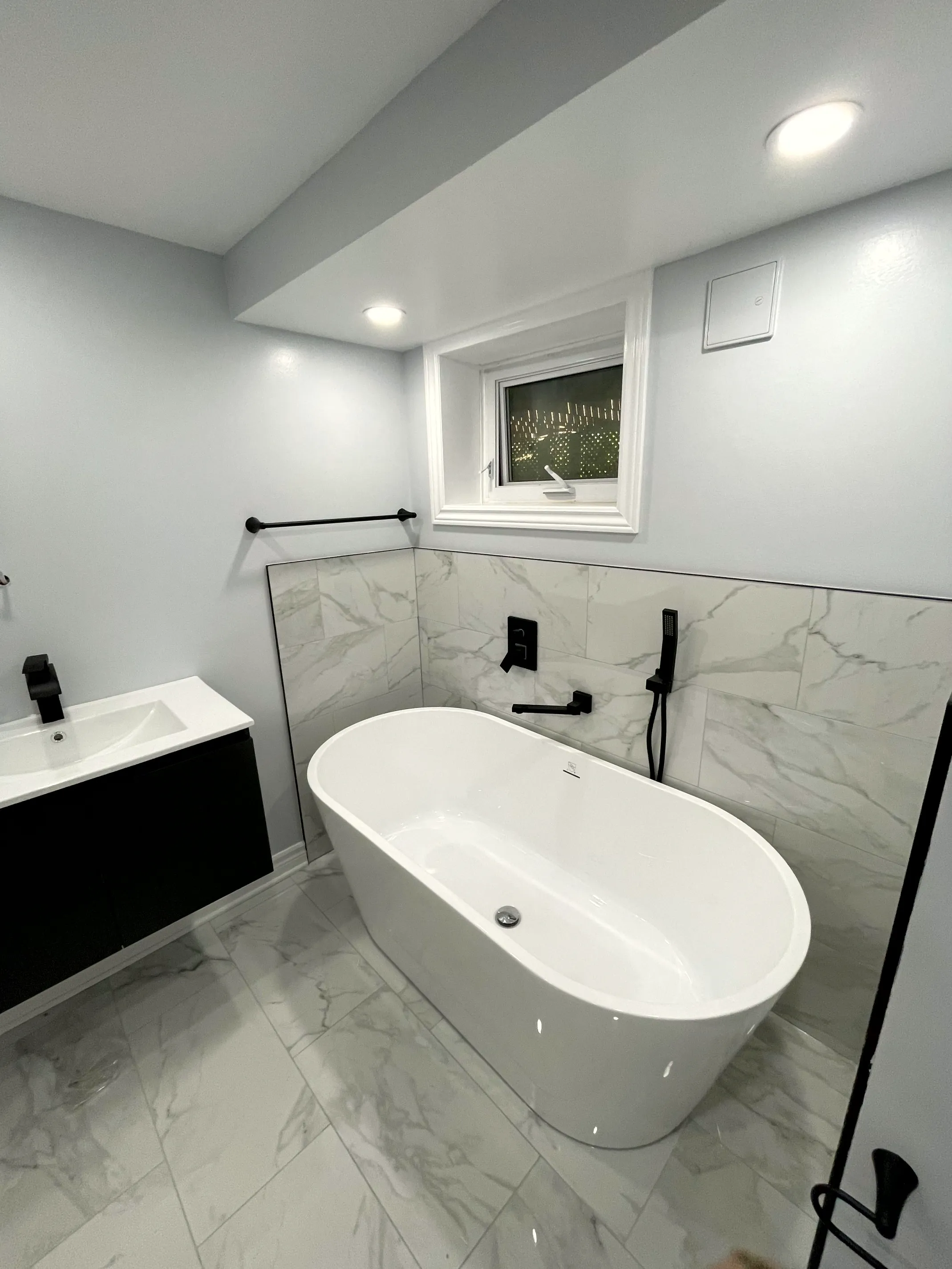 Finished basement bathroom with a freestanding tub, stone-look wall tile, matte black fixtures, and a black floating vanity.