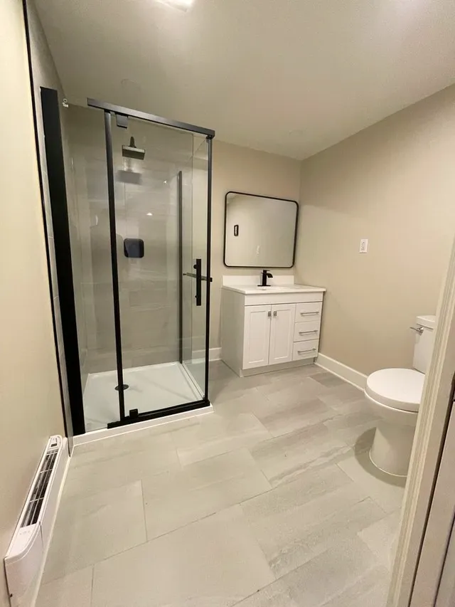 Finished basement bathroom with a black-framed glass corner shower, white vanity, framed mirror, and grey wood-look floor.