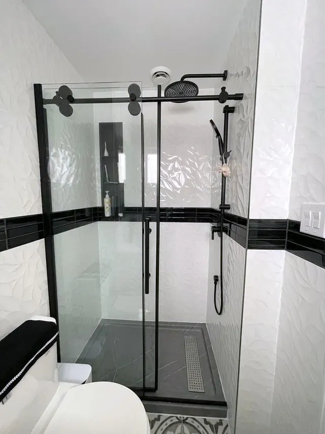 Finished walk-in shower with textured white wall tile, black accent band, sliding glass door, and matte black rainhead and handheld.
