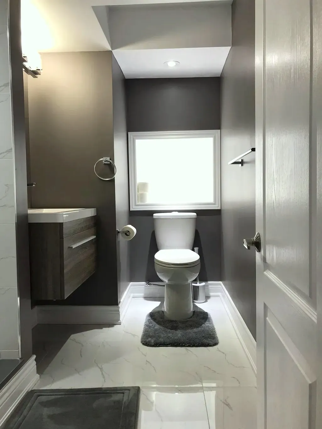 Finished powder room with grey walls, wood-look floating vanity, window above the toilet, and light stone-look floor tile.