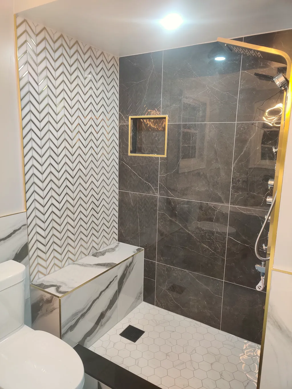 Finished walk-in shower with a chevron-tile accent wall, dark stone-look tile, gold fixtures, a tiled bench, and hex mosaic floor.