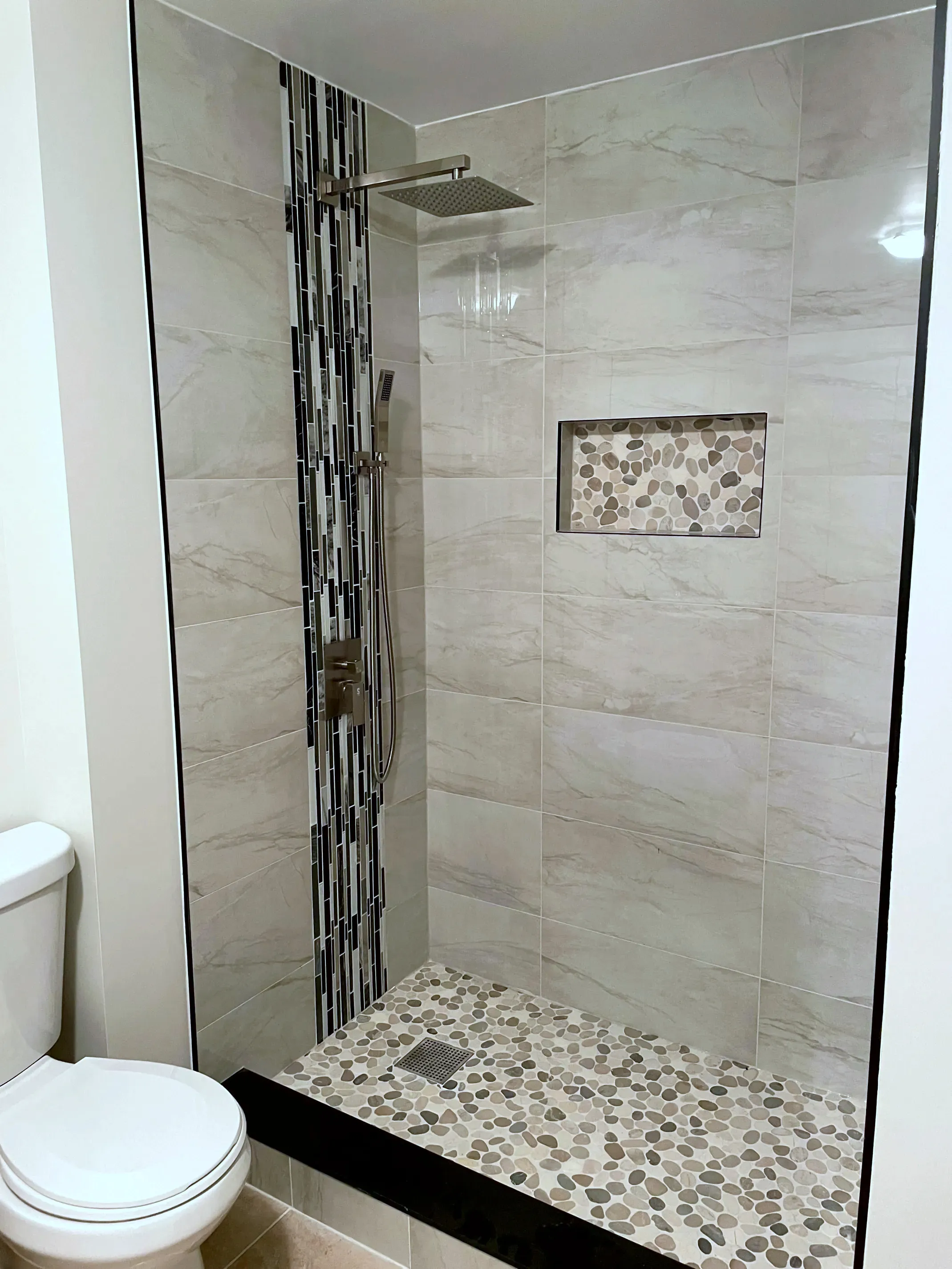 Finished walk-in shower with a vertical glass-mosaic accent strip, chrome rainhead and handheld, and a pebble-mosaic niche and floor.