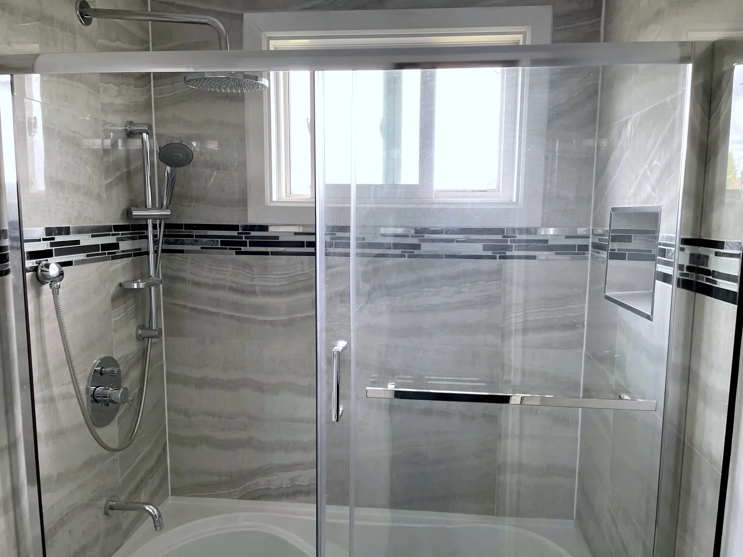 Finished tub-and-shower combo with grey wall tile, a glass-mosaic accent band, a sliding glass door, and chrome rainhead.