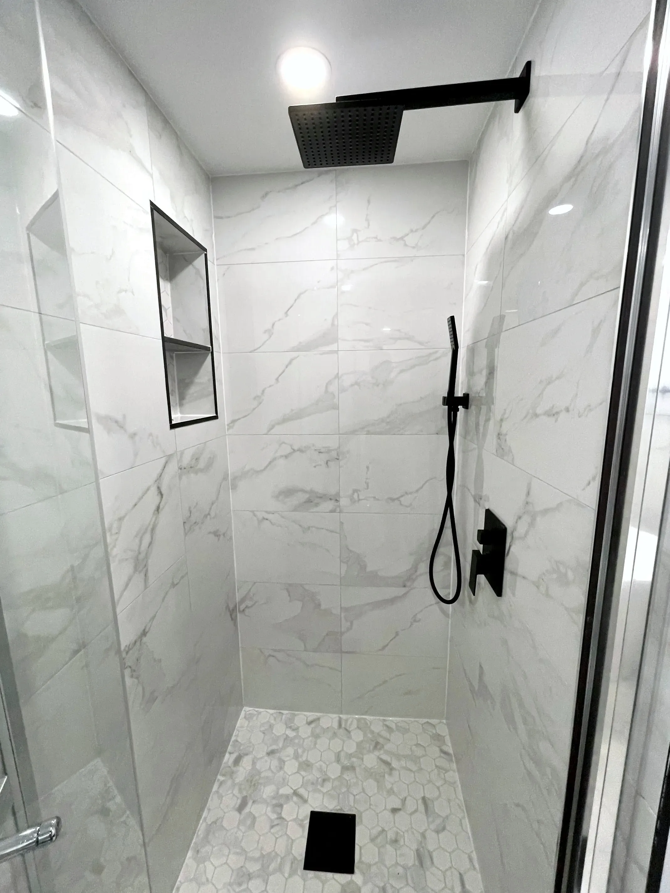 Finished walk-in shower with light stone-look wall tile, a matte-black rainhead and handheld, a recessed niche, and hex mosaic floor.