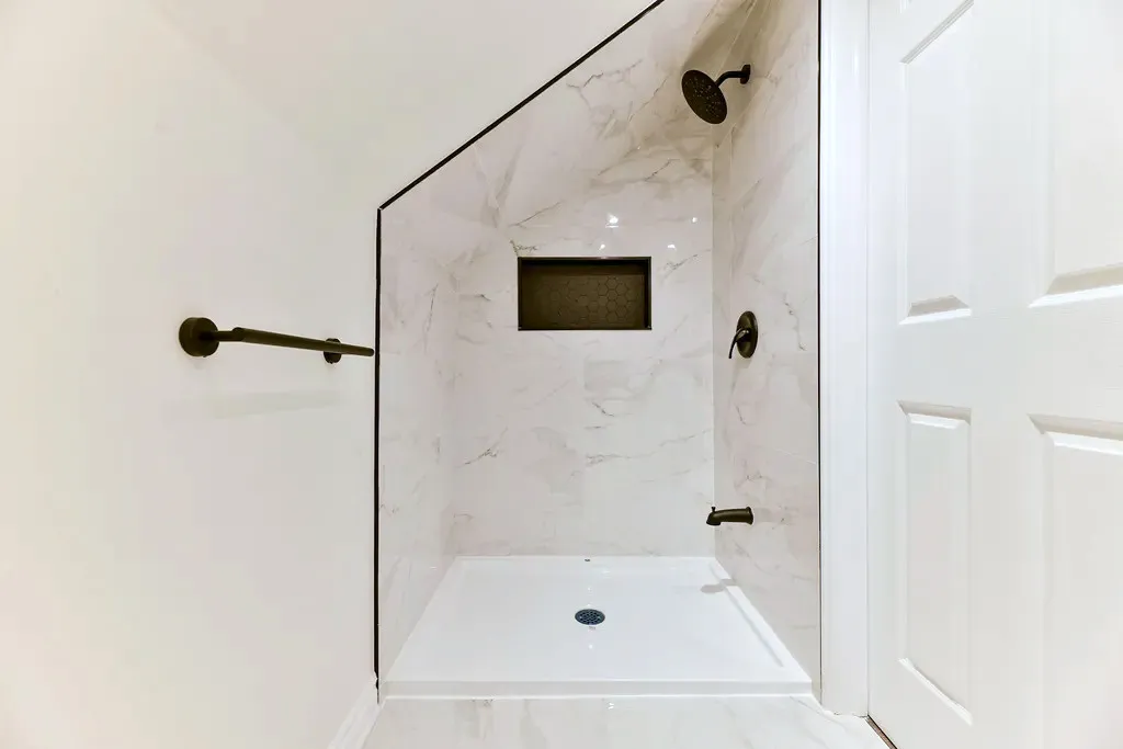 Finished shower under a sloped ceiling with light stone-look wall tile, matte-black fixtures, a recessed niche, and white shower pan.
