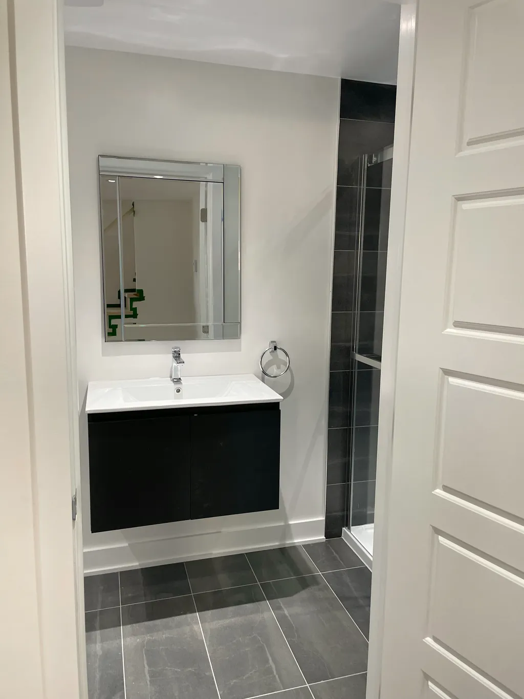 Finished bathroom, black floating vanity, framed mirror, chrome towel ring, glass shower and dark grey tile floor.