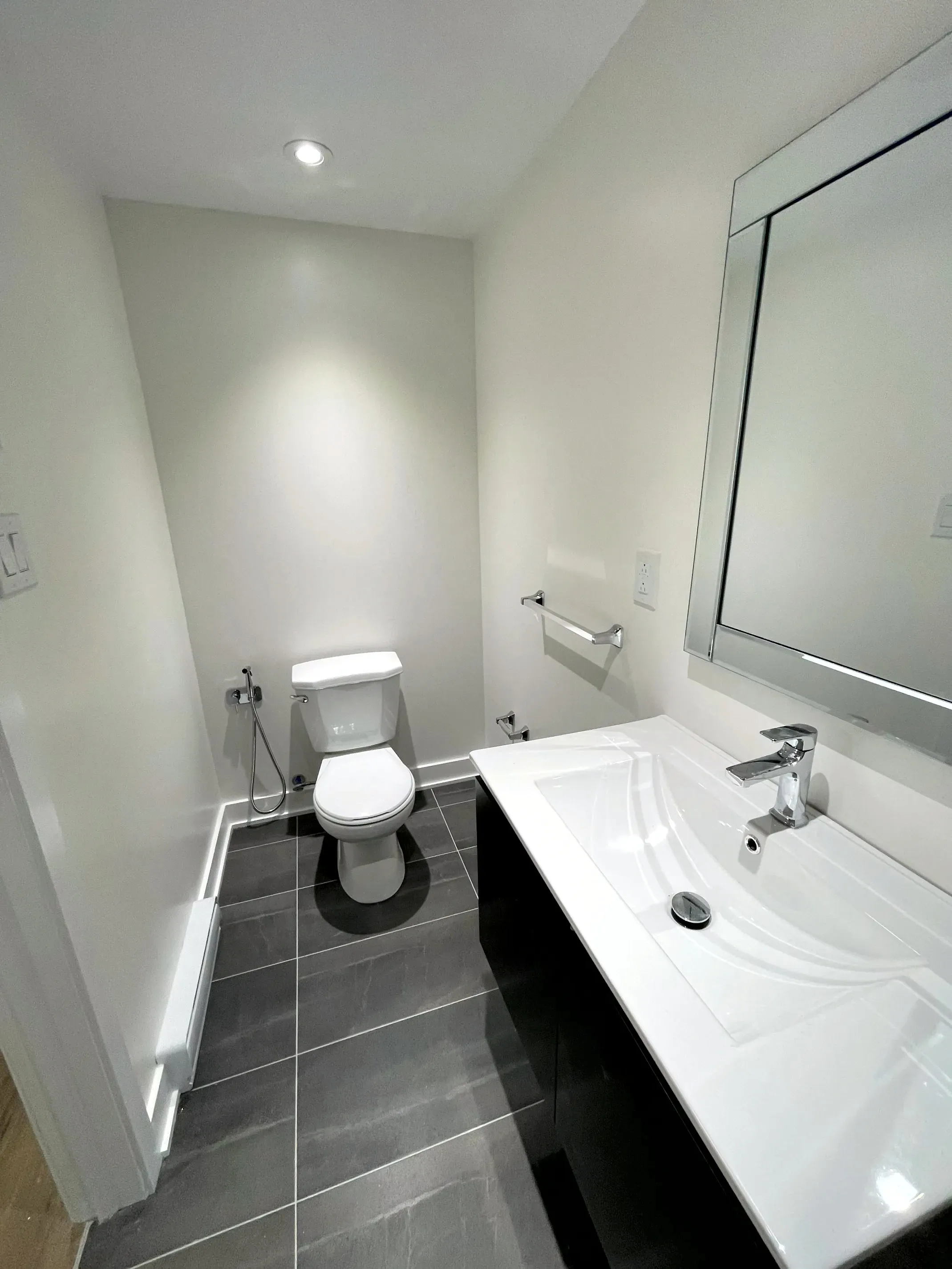 Finished half bath, toilet with hand bidet sprayer, black floating vanity, framed mirror and dark grey floor tile.
