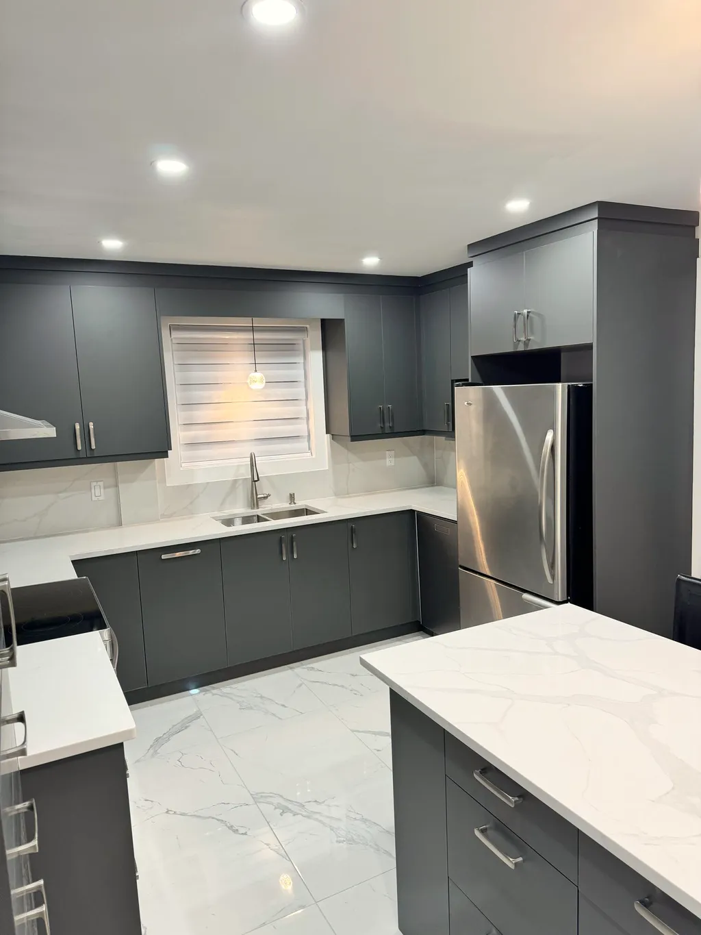 Finished kitchen with dark grey cabinets, white stone-look counters, stainless fridge, zebra-blind window over the sink, and stone-look floor.