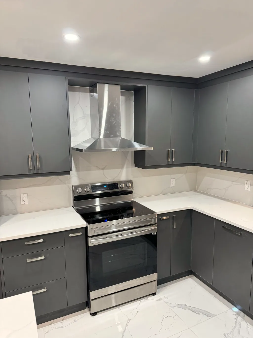 Finished kitchen with charcoal flat-panel cabinets, stainless chimney hood, stainless range, and stone-look counters and backsplash.