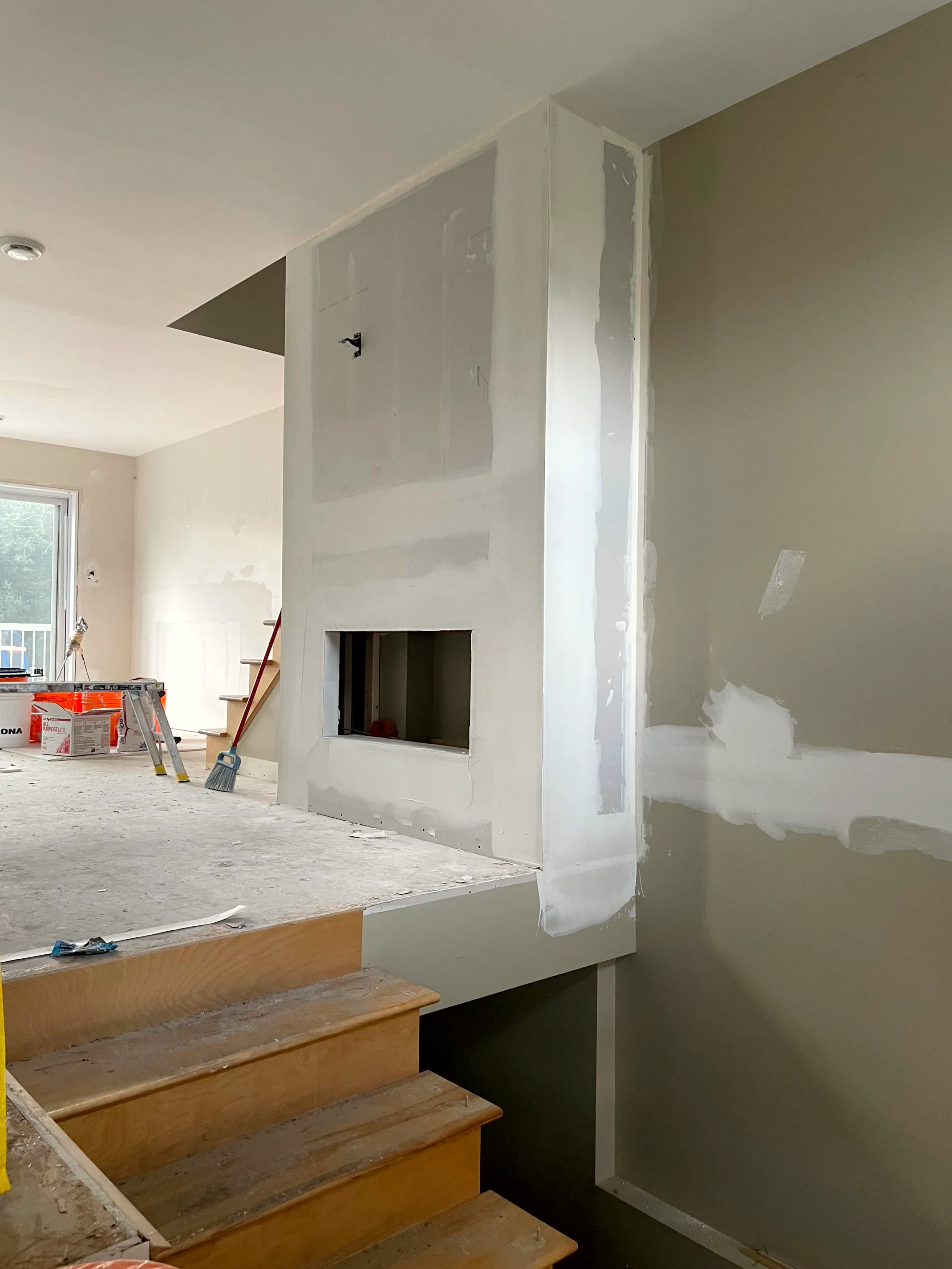 Drywalled fireplace wall with a taped insert opening, beside a wood staircase and floor.