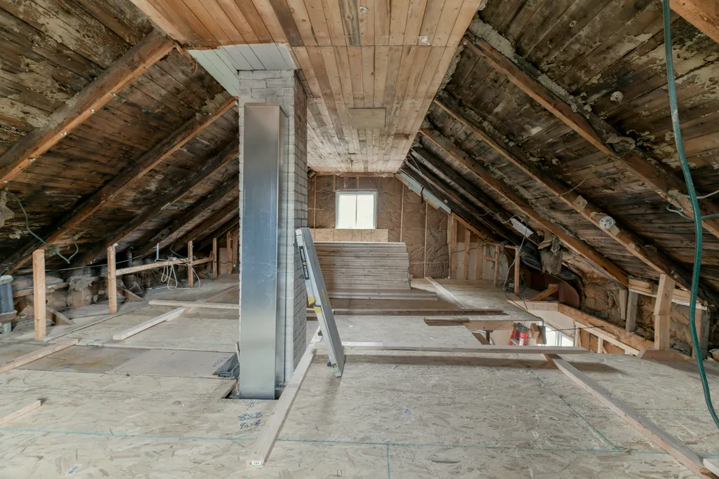 Attic with exposed rafters, new subfloor panels down, a metal chimney chase up the centre and a small window.