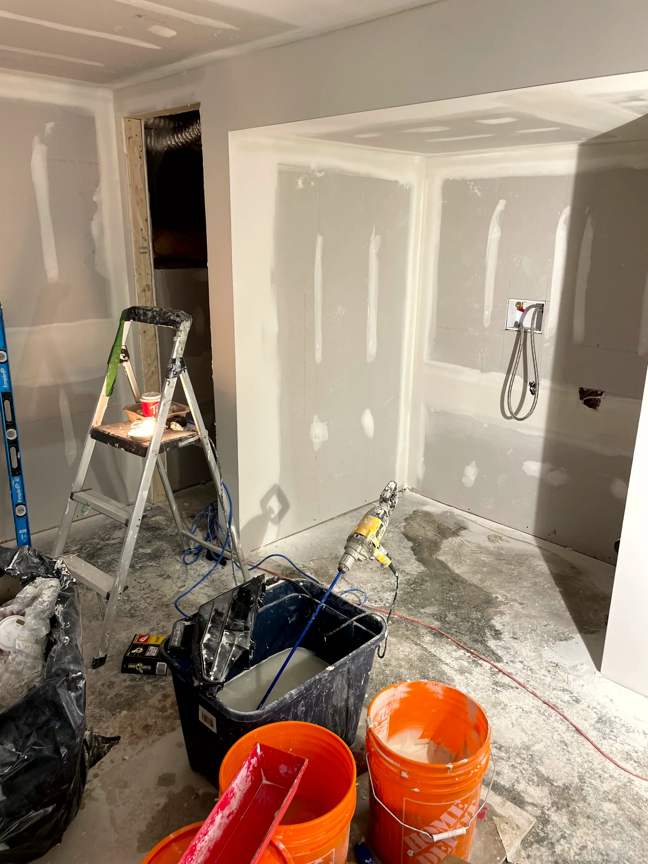 Drywalled bathroom with taped corner walls, a shower valve and handheld set, ladder and buckets on the floor.