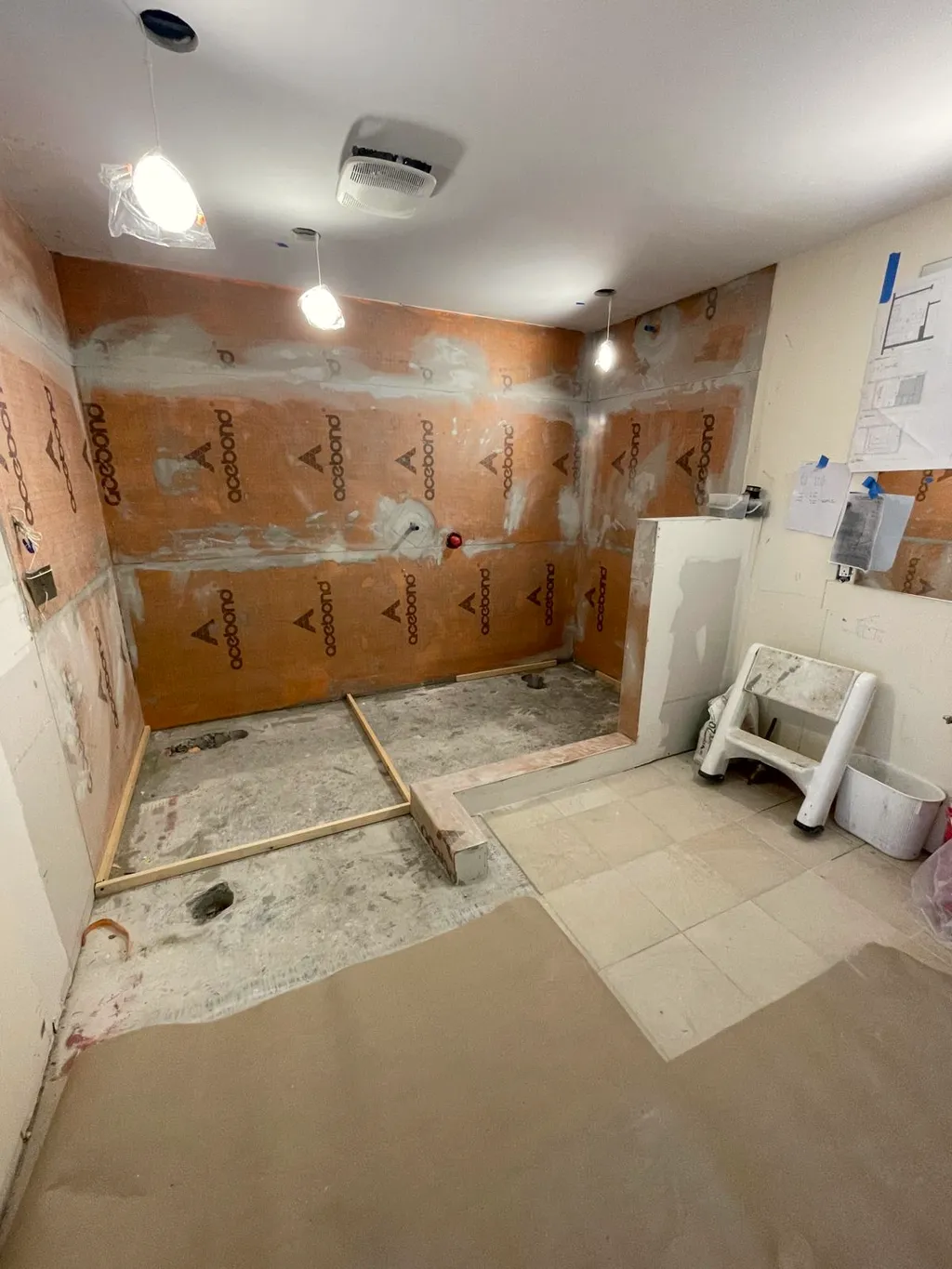 Bathroom with walls clad in board, pendant lights hung, floor tile partly down and a step stool by the wall.