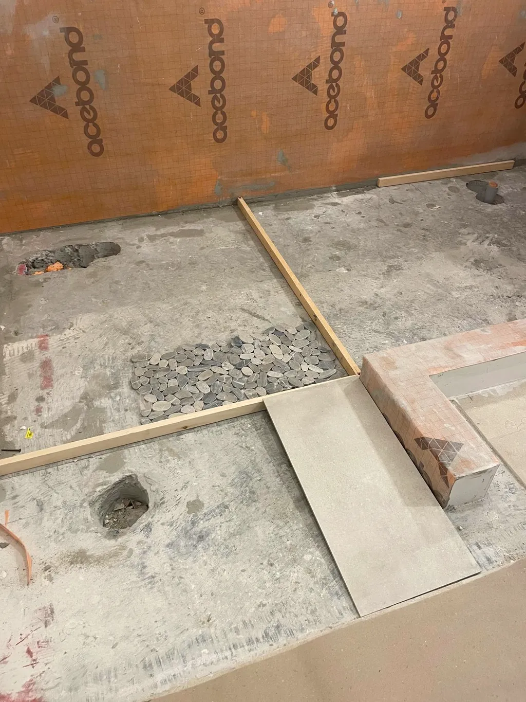 Bathroom floor with a guide batten, a patch of pebble mosaic and a loose tile against a board wall.