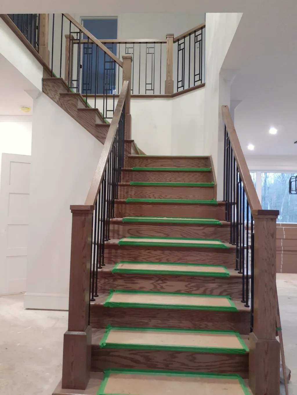 Stained wood staircase with iron balusters, a wood handrail, and treads protected with tape.