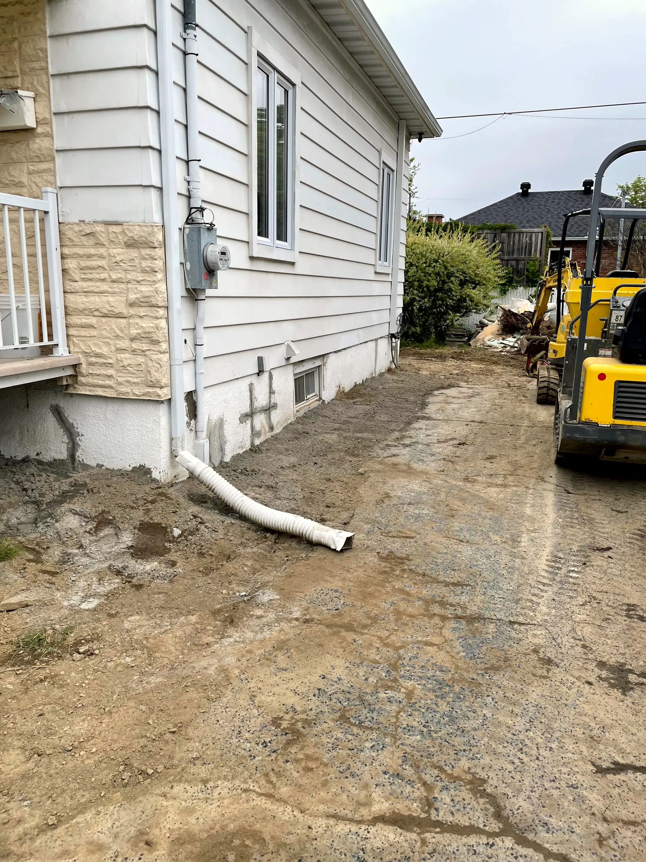 Exterior drain pipe running from the foundation across a graded dirt yard, a mini-excavator parked alongside.