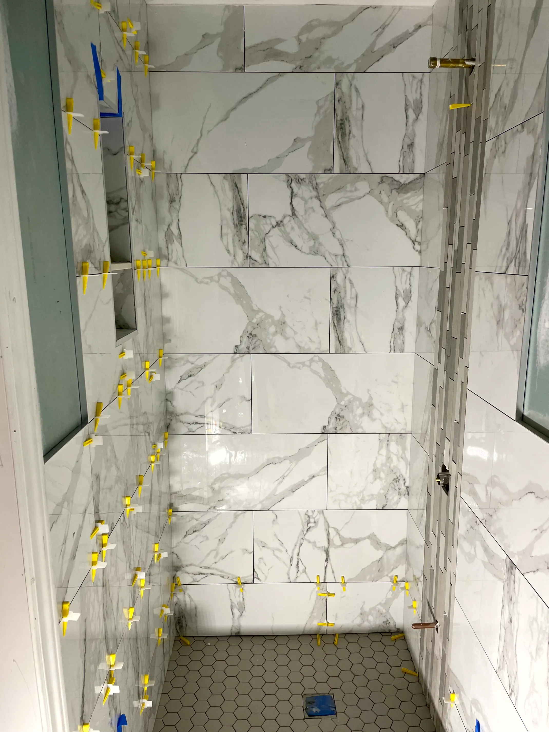 Shower wall tile set with leveling clips, a recessed niche, and a hex mosaic floor below.